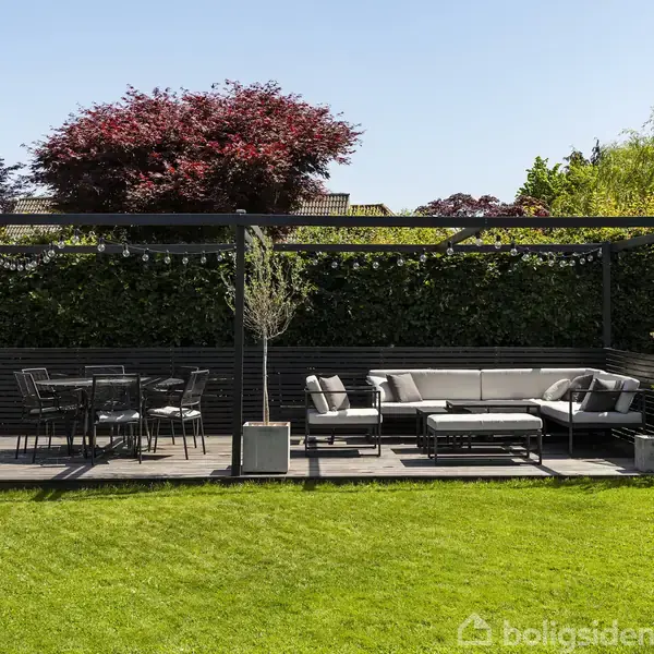 En terrasse med udendørs møblering inkluderer et bord med stole og en sofagruppe. Den står under en pergola omgivet af grønne buske og en velplejet græsplæne på en solrig dag.