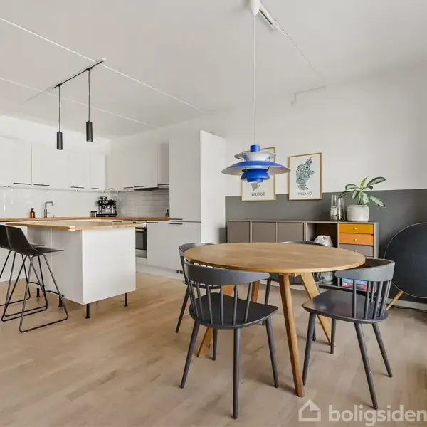 Et åbent køkken-alrum med lyst trægulv, en spisebord med stole og et køkkenbord med barstole. Store vinduer giver udsigt til en altan og bygninger udenfor.