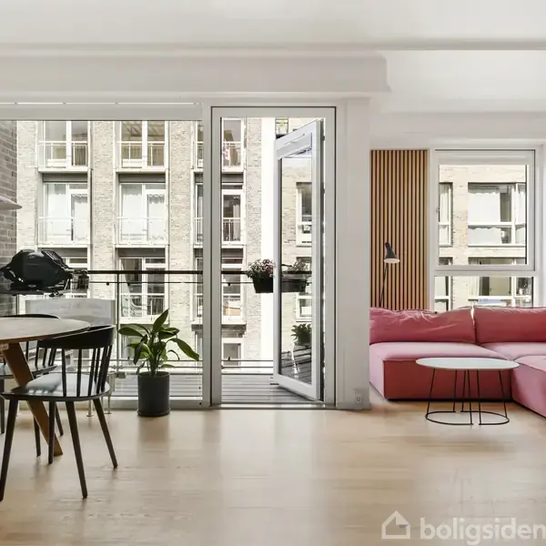 En pink sofa står i en lys stue ved siden af et rundt bord. Spisebord og stole er til venstre, mens en åben dør fører til en balkon med planter.