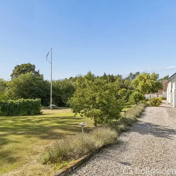 En grusvej fører op til et hvidt hus til højre, omgivet af en frodig have med græsplæne, træer og en flagstang i baggrunden.