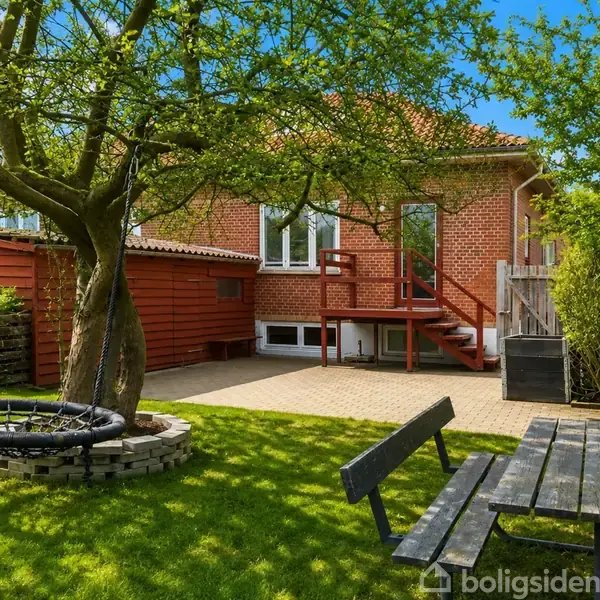 En have med græsplæne hvor et sort gyngestativ hænger fra et træ. I baggrunden står et rødt murstenshus med en terrasse og trappe. En træbænk står i forgrunden.