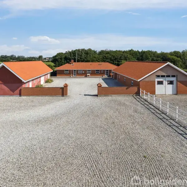 Et rødt murstenshus med orange tegltag står mellem to udhuse. Stenbelagt gårdsplads og grønne marker omkranser ejendommen under en blå himmel.