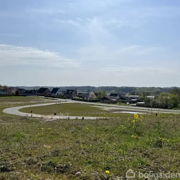 En snoet vej strækker sig gennem et åbent landskab med græs og jord. I baggrunden ses en enkelt bygning og nogle huse. Himlen er klar med spredte skyer.