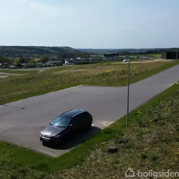 En græsbevokset bakke strækker sig under en blå himmel. Et lille vandhul ses i forgrunden omgivet af sten. Landskabet virker åbent og landligt.