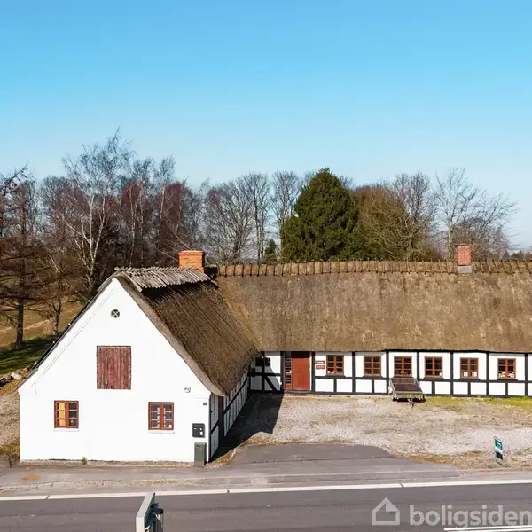 Bindingsværkshus med stråtag står stille ved en vej i et åbent landskab omgivet af træer og blå himmel. Huset har hvide vægge og flere vinduer med brunrammer.
