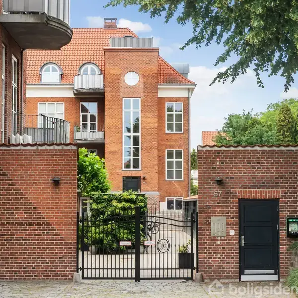 Murstensbygning med rød teglsten; sort jernlåge adskiller gård med grønne planter. Huset har store vinduer og balkon. Nummer 57 fremgår ved indgang. Omgivet af træer og blå himmel.