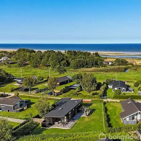 Flere huse står stille omgivet af grønne haver og træer i et kystområde, der strækker sig ud til havet i baggrunden, under en klar blå himmel.