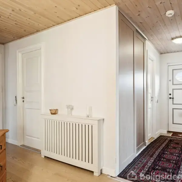 Gang med trægulv, hvidmalede vægge og lyst træloft. Til venstre en kommode med lampe. Døre i midten og for enden af gangen. Tæppe langs gulvet.