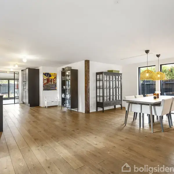 Et lyst, rummeligt køkken-alrum med trægulv. Spisebord med stole står ved vinduer. Skabe og hylder fylder væggen til venstre. Store vinduer giver udsigt til en have.