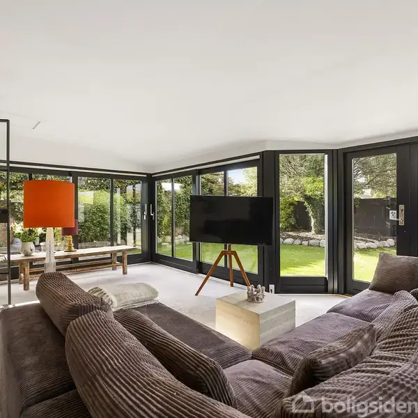 Stor hjørnesofa i brun farve vender mod et fladskærms-tv på stativ. Rummet har store vinduer, der viser en grøn have udenfor. En orange lampe står til venstre.