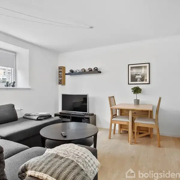 Grå sofa med puder står i stuehjørne. Rundt sofabord foran. Fjernsyn ved siden. Lille spisebord med to stole og plante. Hylde med småbiler. Billede på væggen.