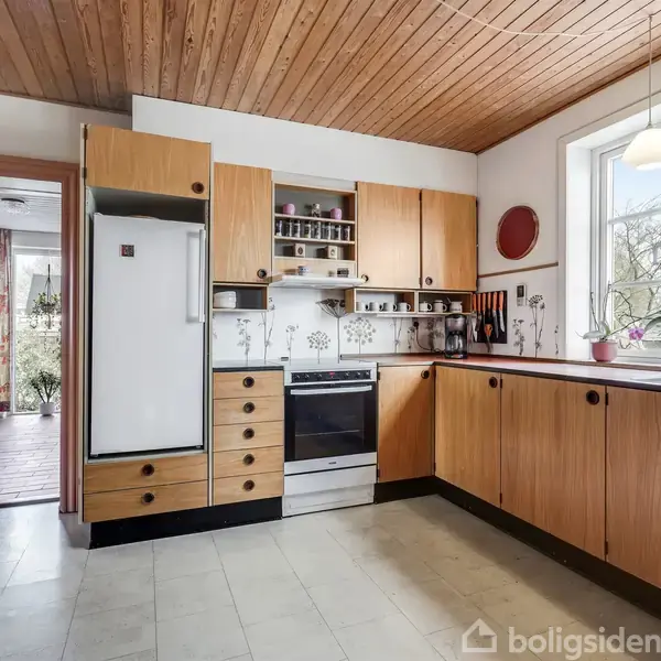 Køkken med træskabe, hvidt køleskab og komfur; flere krydderier på hylde. Vinduet har udsigt til træer. Til venstre ses en åbning til en anden stue.