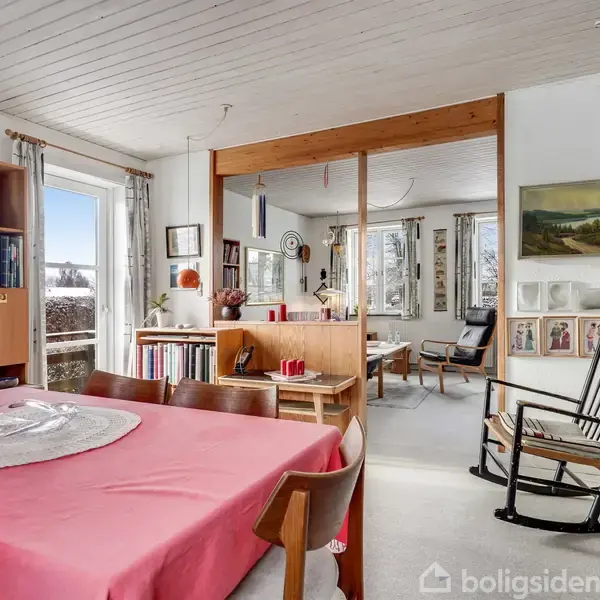 Spisebord dækket med en lyserød dug står centralt, omgivet af stole. I baggrunden ses en reol med bøger og indrammede billeder på væggen, i et lyst rum med vindue.