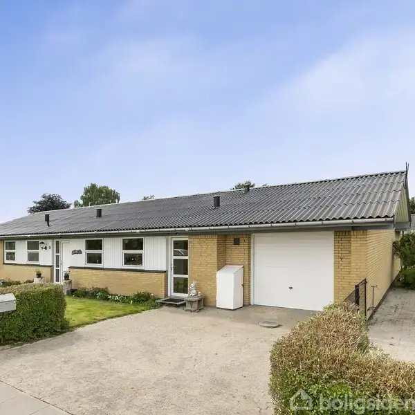 Et parcelhus med gul murstensfacade og gråt tag står i en forstadsomgivelse. En grøn græsplæne og hække omkranser huset. En garage er integreret i siden af bygningen.