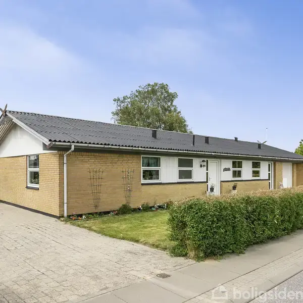 Et gult murstenshus med mørkt tag står ved en hæk på en asfalteret vej. Huset har en flisebelagt indkørsel og en velplejet græsplæne.