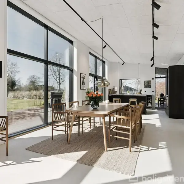 Spisebord med stole står i et lyst, moderne rum med store vinduer. Der er kunst på væggene og en vase med blomster på bordet. Afslappet, minimalistisk indretning. Ingen tekst.