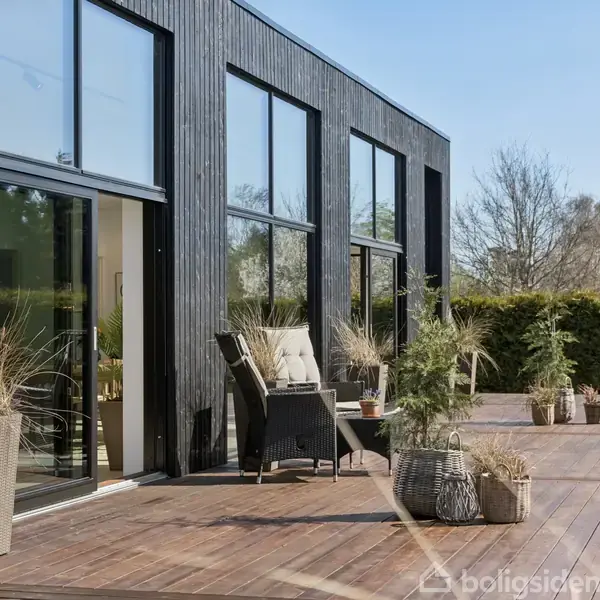 Moderne træhus med store vinduer står på en solrig terrasse. Potteplanter placeret langs kanten skaber en hyggelig stemning. Træer og buske grænser op til området.