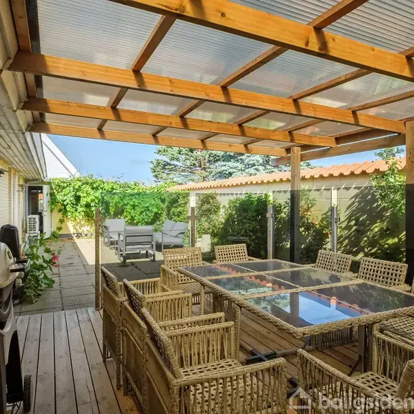 Terrasse med gennemsigtigt tag, bord og stole i kurvemateriale står midt i rummet. Gasgrill til venstre. Omringet af grønne planter, med udsigt til haven.