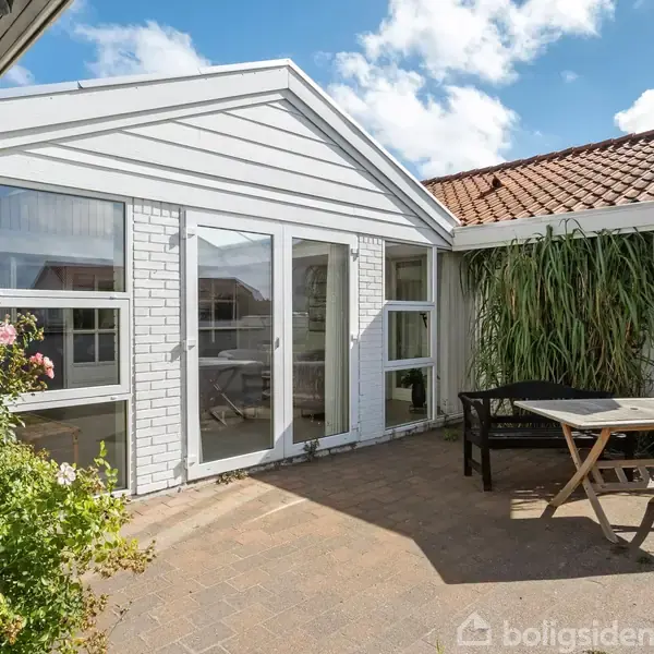 Hvidmalet hus med store glasdøre åbner ud mod en solrig terrasse. Et bord med stole står ved siden af en væg med grøn beplantning. Blå himmel i baggrunden.
