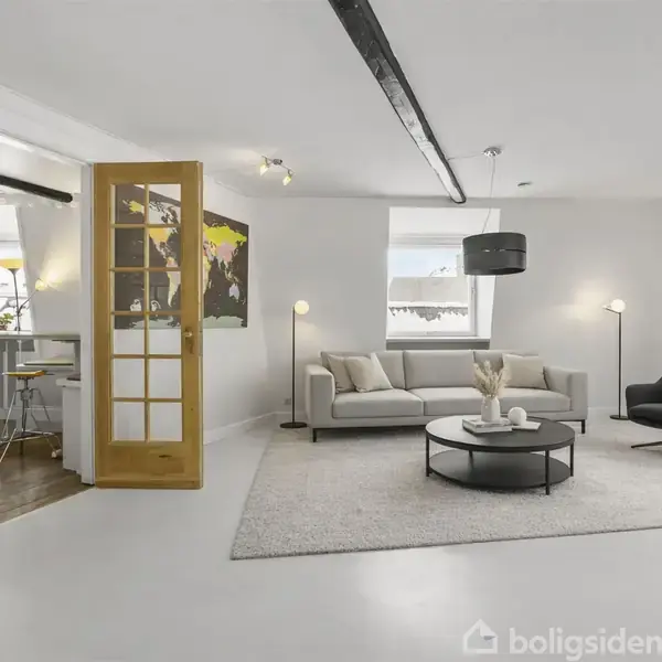 Lys stue med sofa og lænestol, placeret på tæppe i moderne indretning. Rundt sofabord med dekorationer. Åben dør viser spiseområde. Væglampe skaber hyggelig atmosfære.