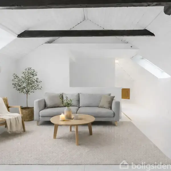 En stue med en grå sofa og et rundt træbord foran. En lænestol med et tæppe står ved siden af. Et lille træ står ved sofaen. Rummet har lyse træbjælker og hvide vægge.