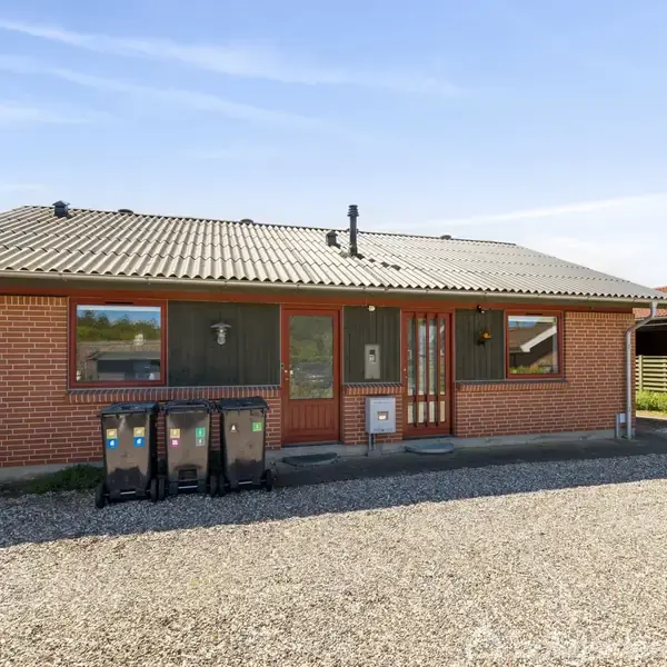 Étplans murstenshus med sort tag, placeret i en have med græsplæne, flisebelagt terrasse og en hævet plantagekasse. En parasol står tæt ved terrassen i det udendørs miljø.
