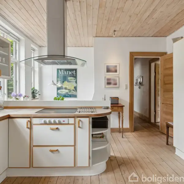 Et lyst køkken med hvidt og trædekor, hvor der står et komfur med en emhætte. Store vinduer giver naturligt lys. En plakat med teksten "Manet" hænger på væggen.