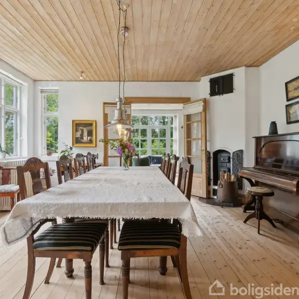 Spisestue med et langt bord, der er dækket med en dug og omgivet af stole. Et klaver står mod væggen ved siden af en brændeovn. Store vinduer og parketgulv dominerer rummet.