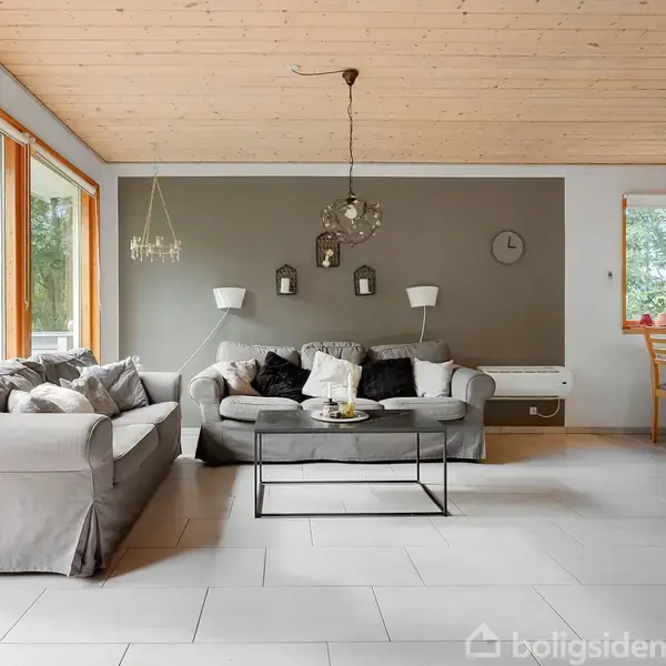 Grå sofaer står i en stue med lyse flisegulve og træloft. En grå væg med vægure og lamper. Store vinduer giver udsigt til træer. Spisebord ses til højre.