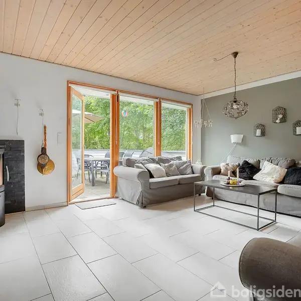 Brændeovn opvarmer stuen, mens grå sofaer omkranser sofabordet. Store vinduer åbner ud mod en terrasse og grønne omgivelser, med en lys træloft i rummet.