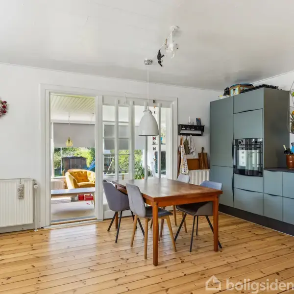 Et spisebord med stole står i et moderne køkken med trægulv og grå skabe. En dør fører til en lys stue med planter og lysindfald fra vinduer.