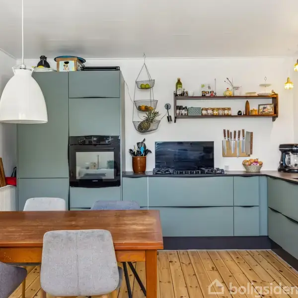 Et moderne køkken med grå skabe, integreret ovn og komfur. På trægulvet står et spisebord med grå stole. Hylder med krydderier og lamper hænger på de hvide vægge.