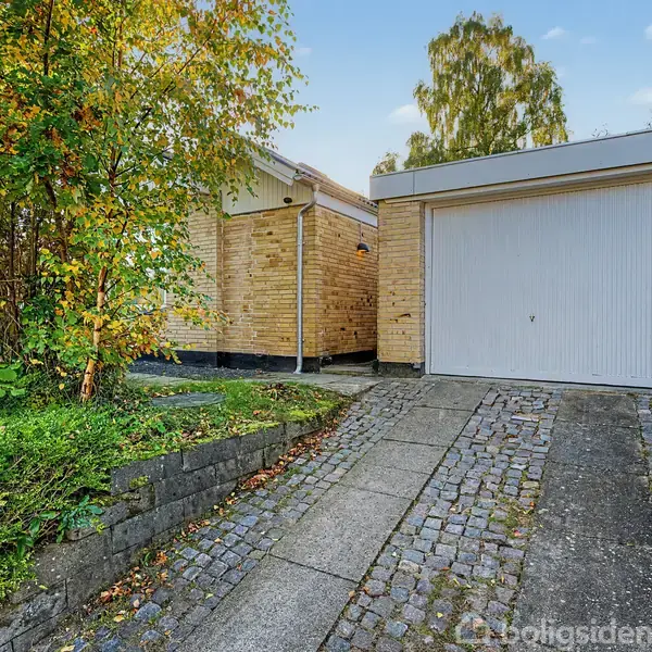 Garageport i hvidt, lukket foran en murstensbygning. Flisebelagt indkørsel skråner opad, omgivet af grønne planter og træer under en blå himmel.