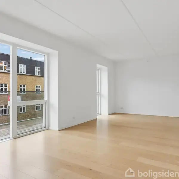 Stue med lysegrå vægge og lyst trægulv, set fra et hjørne. To store glasdøre fører til en balkon. Udenfor ses murstensbygninger og himmel.