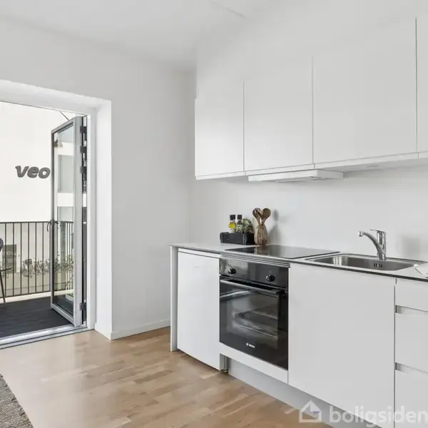 Et moderne køkken med hvide skabe indeholder en indbygget ovn og en vask. En dør står åben til en balkon med stole. Udenfor ses en bygning med teksten "Veo".