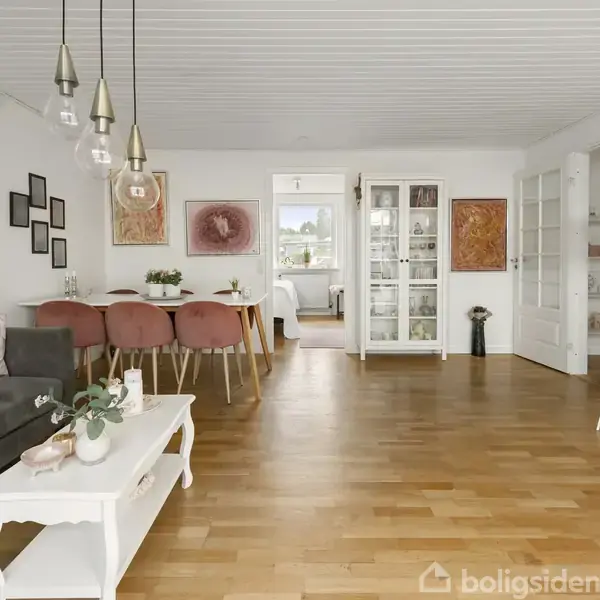En spisestue med seks lyserøde stole omkring et træbord, omgivet af kunst på væggene. I baggrunden ses en åbning til et værelse med vindue og udsigt.