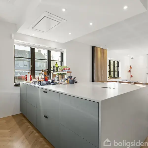 Køkkenø med grå skabsfronter står centralt, omgivet af lysindfald fra store vinduer. Rummet er åbent og lyst med trægulve og moderne indretning. Ingen synlig tekst.