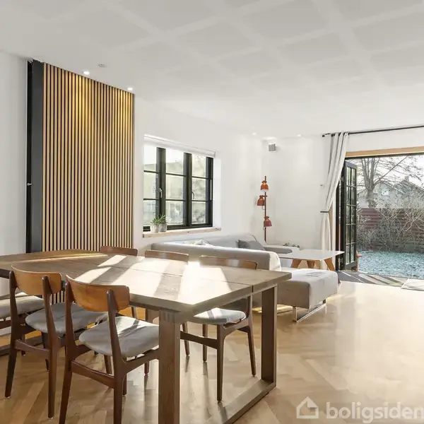 Spisebord med fire stole står i en lys stue. Store vinduer og en terrassedør åbner ud mod en have. Hylder med bøger er på højre side.