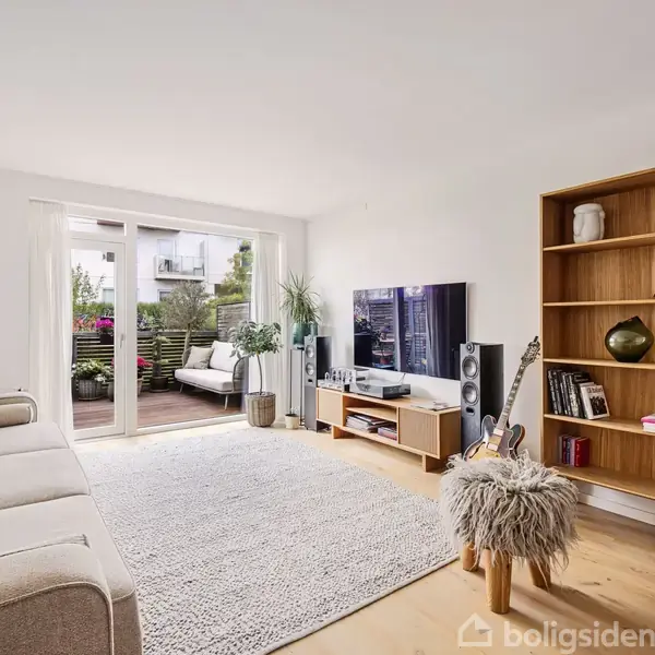 Stue med grå sofa ved siden af en reol. Fladskærms-tv og højttalere står på TV-bord. Store vinduer åbner ud til en terrasse med planter. Tæppe dækker trægulvet.