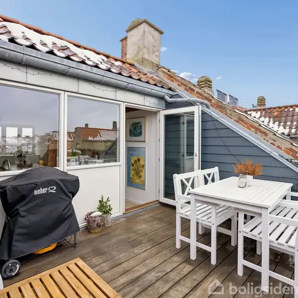 Terrassemøbler står på en altan med trægulv, omgivet af ældre huse med røde tagsten. En grill med overtræk er til venstre, og døren fører ind i huset.