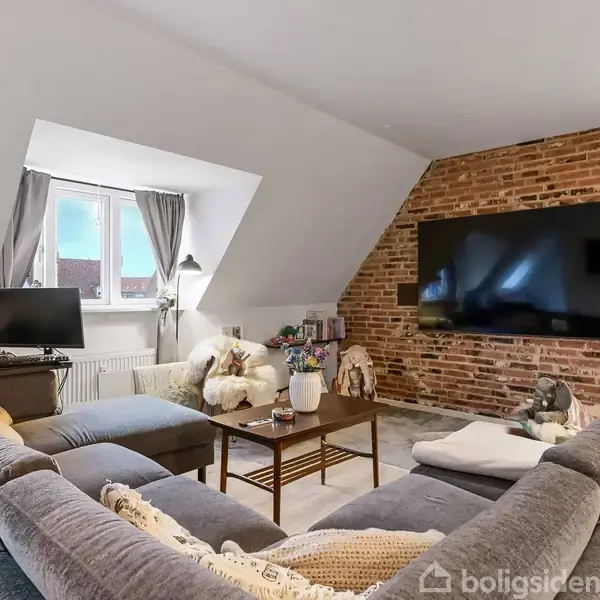 Stor grå hjørnesofa står foran en murstensvæg med stor fladskærm. Et sofabord med planter og dekorationer står i midten. Vindue med grå gardiner ses i baggrunden.