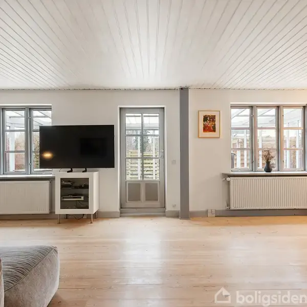 Sofa står i stuen op ad væggen med en plakat. Et rundt bord foran sofaen. I baggrunden en brændeovn ved siden af en vitrineskab. Lyst trægulv og vinduer.