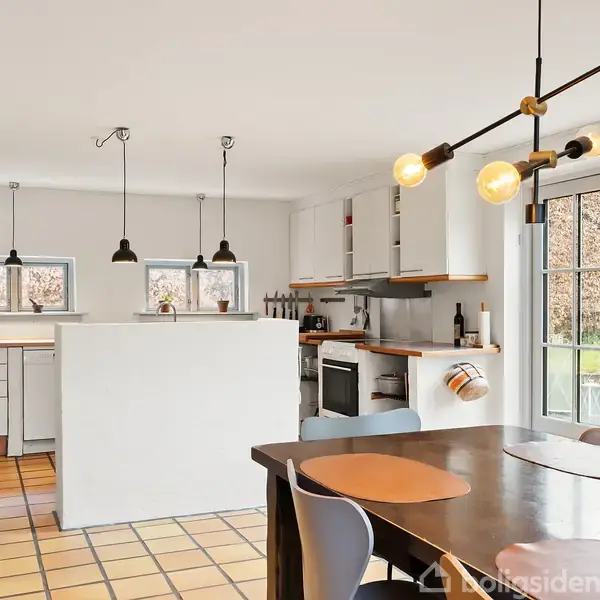 Stue med lys træparket og hvidmalet loft, placeret mellem store vinduer. Et TV står på et hvidt møbel ved siden af en dør. En brun kasse står til højre.