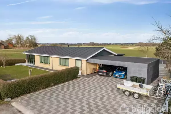 En moderne, rektangulær etplansvilla med garage ligger i en landlig omgivelser. Biler er parkeret i carporten, og en trailer står på en mønstret indkørsel.