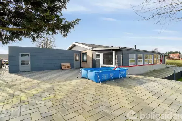 En moderne bungalow med fladt tag står stille på en stor flisebelagt terrasse. Et blåt, oppusteligt badebassin er placeret foran huset. I baggrunden ses et naturområde.