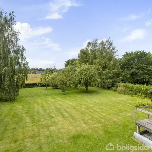 En velplejet, grøn græsplæne med enkelte træer står omkranset af buske og tæt løv. Til højre ses en træterrasse ved siden af et lille rødt skur. Himmel med spredte skyer.