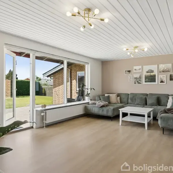 Stor, lys stue med en grøn plante i forgrunden. En stor grå sofa står langs væggen med billeder over den. Et sofabord foran sofaen. Udsigt til en have gennem store vinduer.