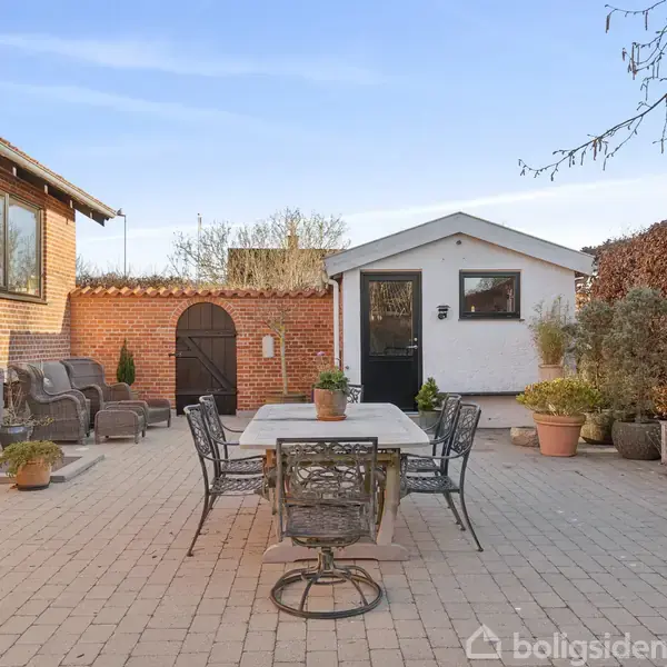 Terrasse med spisebord og stole i metal, omgivet af blomsterkrukker og bænke. Murstensbygning til venstre og hvid haveskur foran, flisebelagt område i solskin.