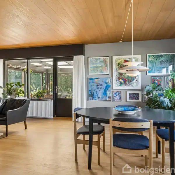 Rundt spisebord i moderne spisestue med trestole, omgivet af kunst på væggene og grønne planter ved store vinduer. I baggrunden ses sort lænestol nær glasdør til udestue.