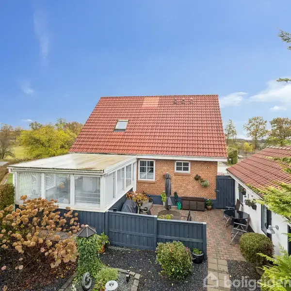 Et rødt murstenshus med rødt tag ses fra haven, hvor der er en flisebelagt terrasse med møbler. Omgivet af grønne træer og buske i efterårsfarver.